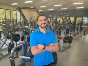 a man in a blue shirt stands in a gym in front of a room full of weightlifting machines.