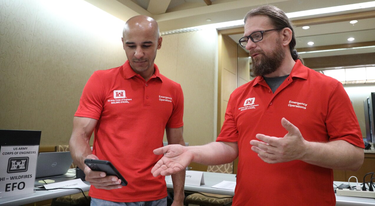 Two men wearing red shirts with U.S. Army Corps of Engineers logos look at a smartphone.