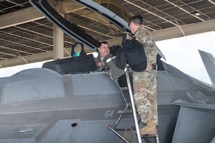 Crew chief standing on ladder getting bag from pilot in F-22 cockpit.