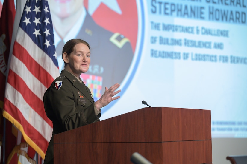 A female uniformed service member stands behind a lectern in front of a presentation.