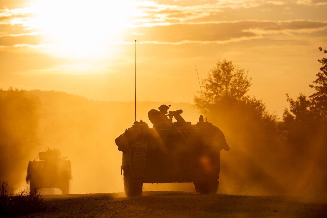 Soldiers riding in various armored vehicles roll through a training village at dusk.