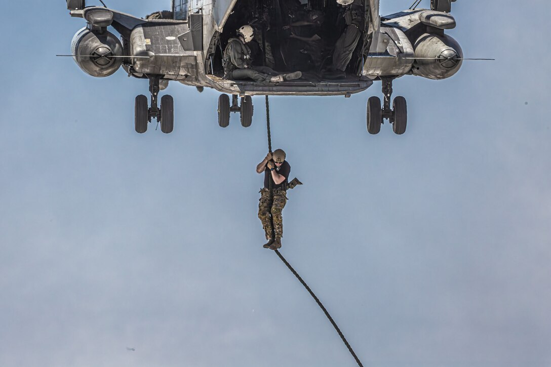 A soldier hangs on to a rope below a flying helicopter.