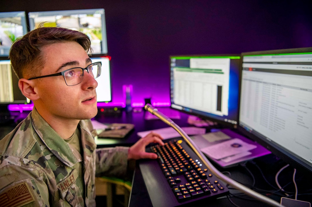 An airman sits at a desk in front of computer monitors.