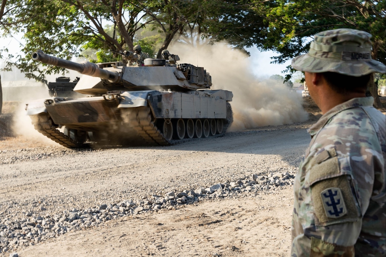 A soldier watches a tank drive by on a dirt road.