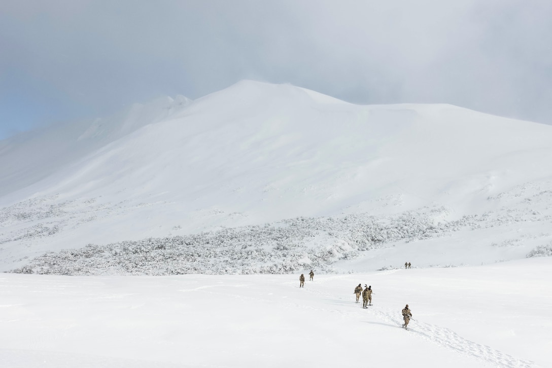 Several service members are seen from a distance traveling against snowy, mountainous terrain.