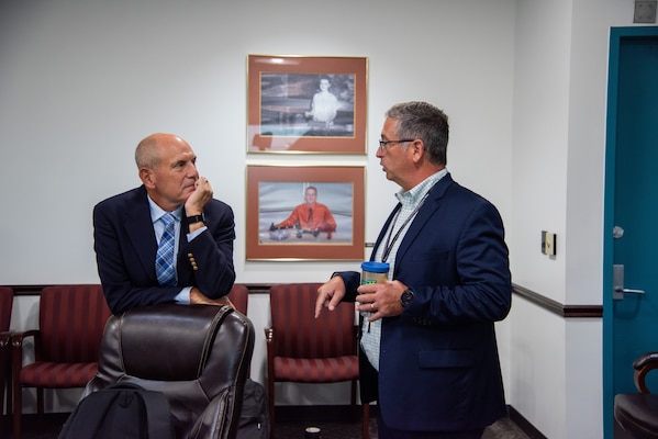 Two men chat at a conference room table. Both are in business attire. One is bald and the other has grey hair and glasses.