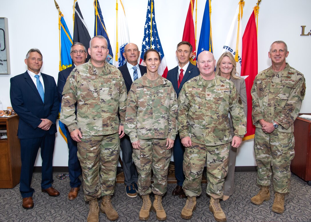 A large group of people in military uniforms and business attire stand in front of a group of military flags. All are smiling.