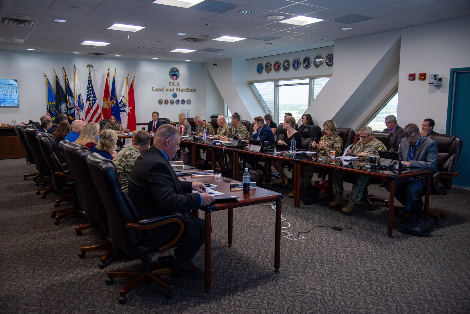 A group of people sit in a conference room at a large table. Some in military uniforms and some in business attire.