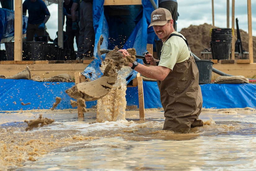 An airman wearing a waterproof suit throws mud with a shovel while standing in a pond with a wooden structure in the background.