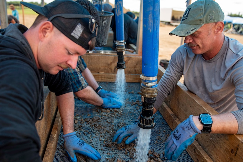 Two soldiers wear gloves while handling dirt on a wet-screening device.