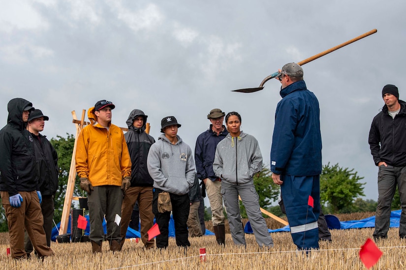 A man holds a shovel as he talks to a group of people standing in a field.