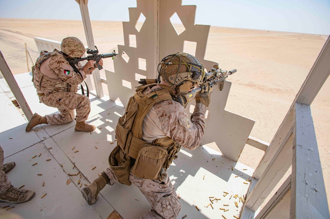 A Marine fires a weapon from behind a barrier next to a foreign service member also firing a weapon.