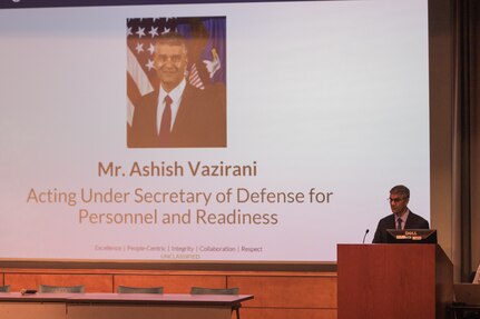 A man in a suit stands on a stage behind a lectern.