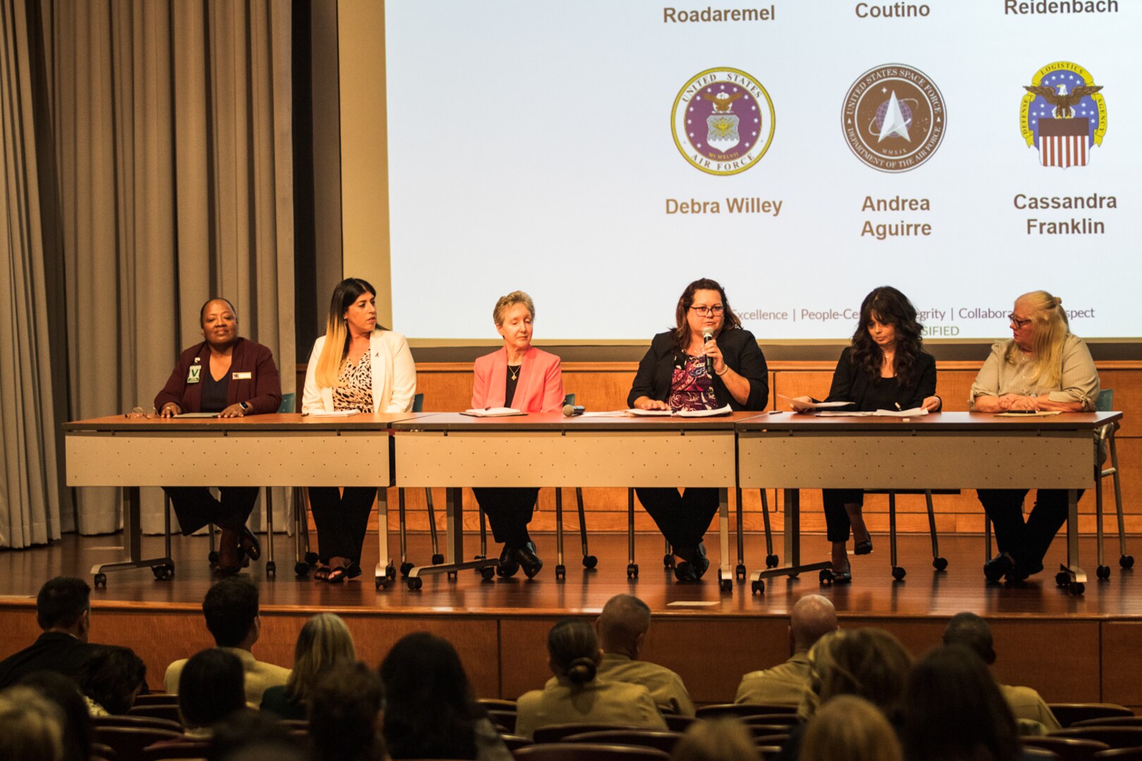 One woman holds a microphone while women are seated to her right and left at table on a stage facing a crowd.