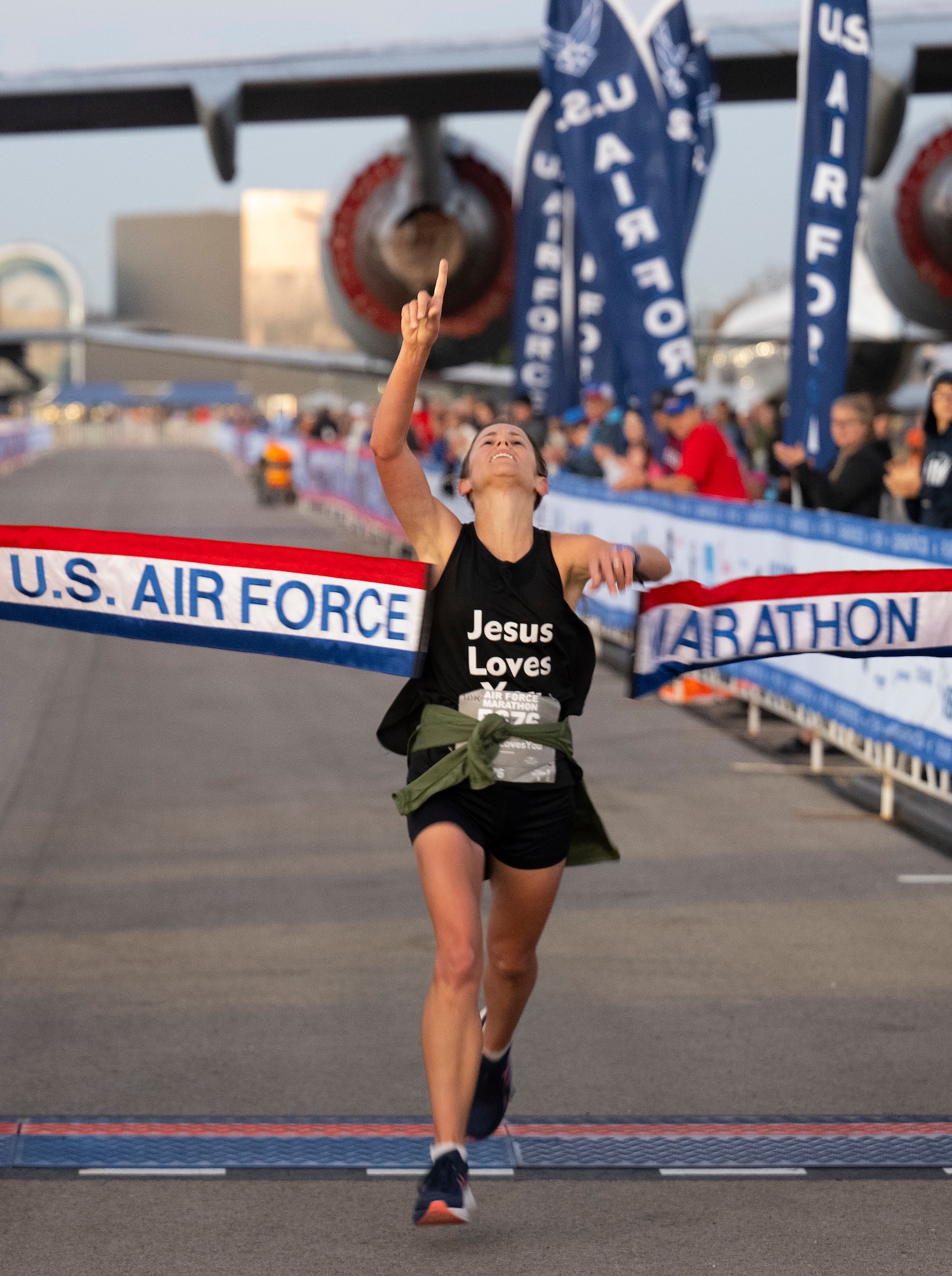 A woman in black athletic clothes points to the sky as she breaks a red, white and blue banner with "US Air Force Marathon" on it