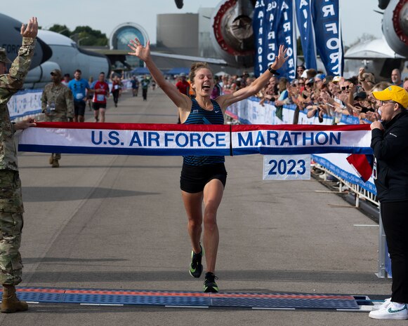 Woman raises her arms as she nears the banner held by a man in uniform and a woman in civilian clothes.