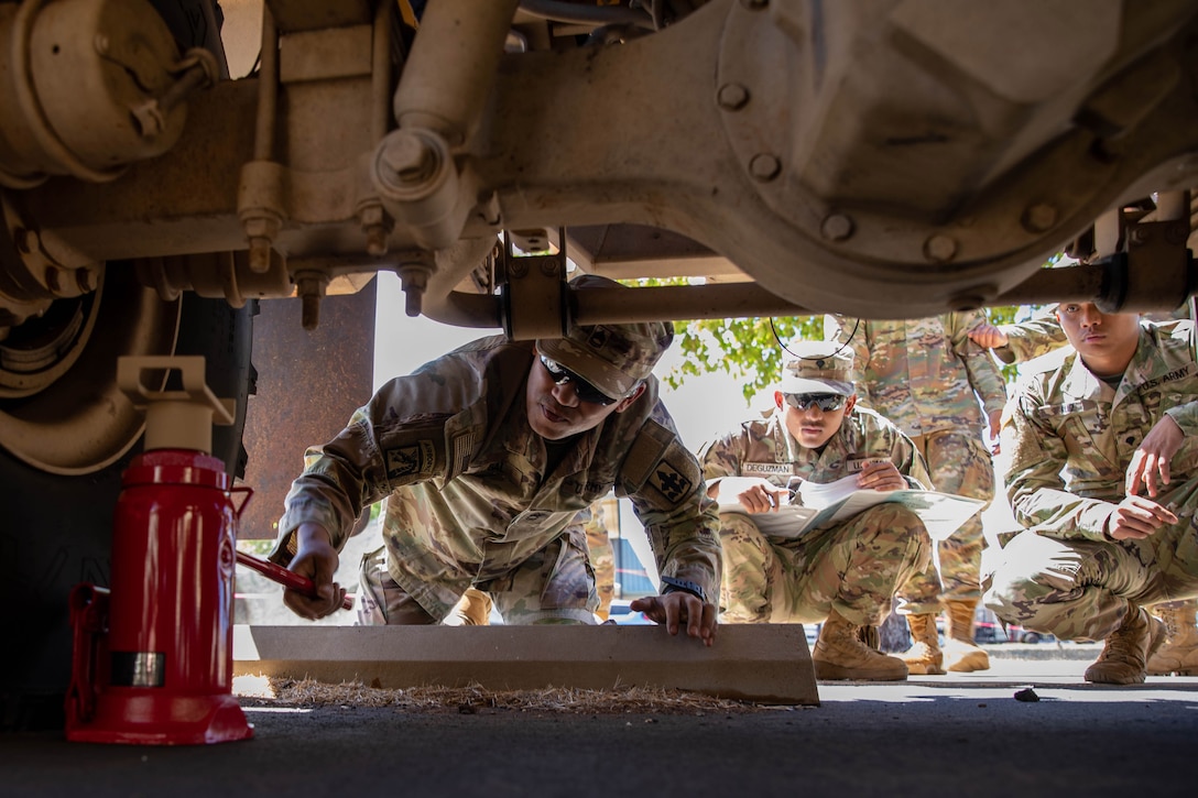 A soldier on his knees under a vehicle holds a metal rod.