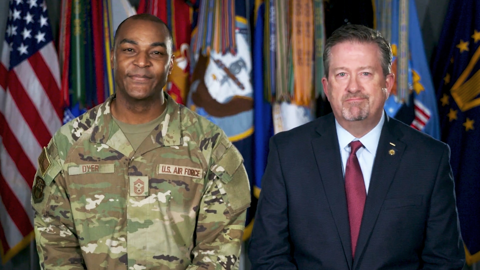 Two men - one in a suit and one in camo uniform - sit in front of a row of flags