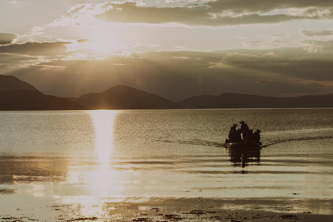 Service members in silhouette sail in a small craft illuminated by low sunlight.