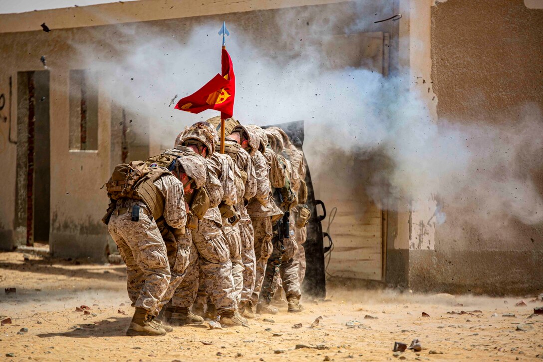 Marines stand huddled together in a line behind a protective shield as an explosive detonates.