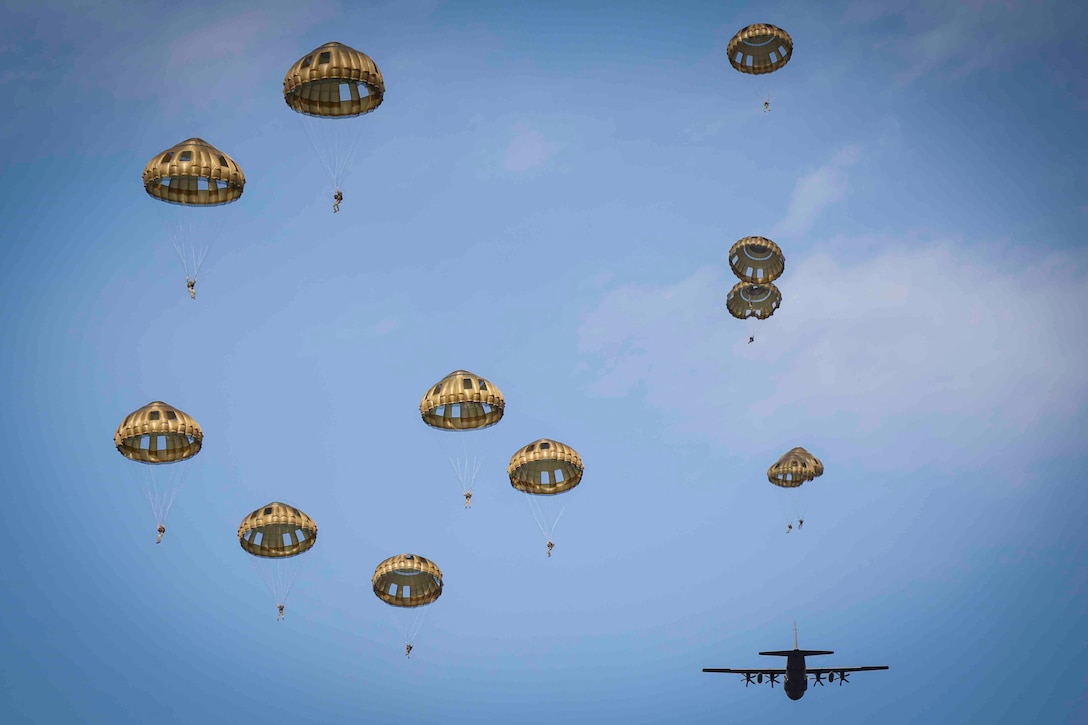 U.S. and foreign service members descend in the sky wearing parachutes as a plane flies in the distance.