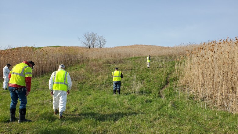 Personnel in protective clothing survey a grass-covered hill.
