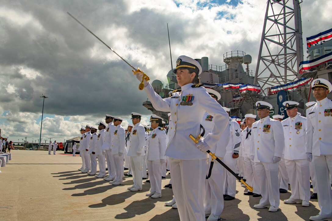 A sailor holds a sword in the air while standing in front of fellow sailors standing in formation with a ship in the background.