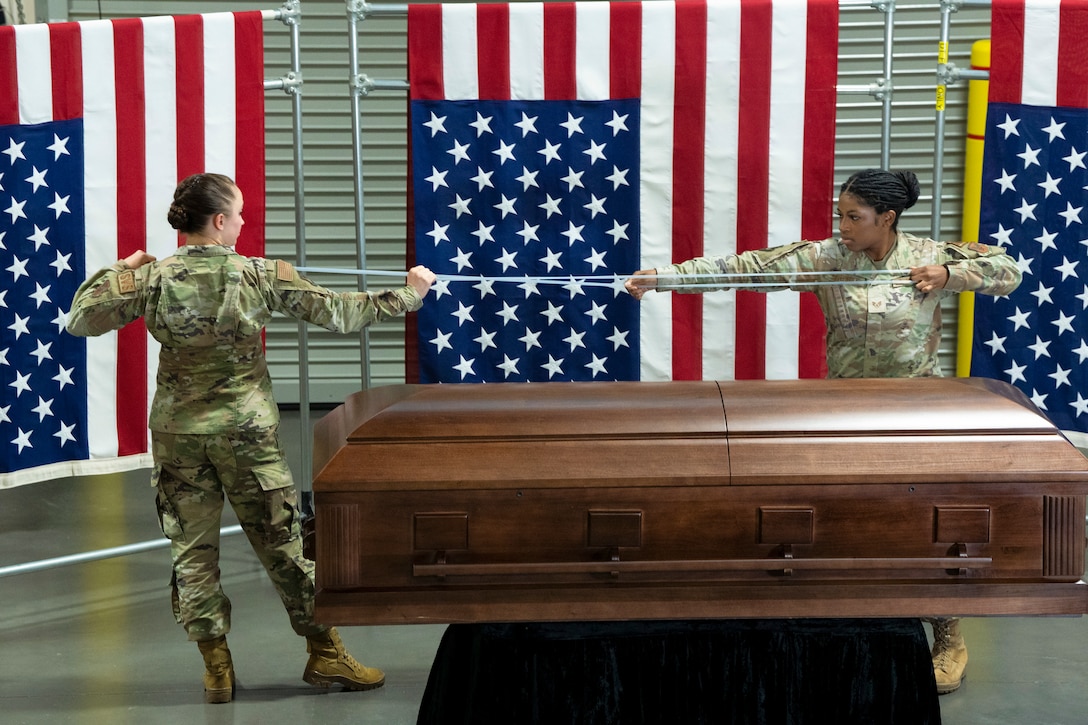 Two airmen stand on opposite sides to stretch a band with three American flags hanging in the background and a casket in the foreground.