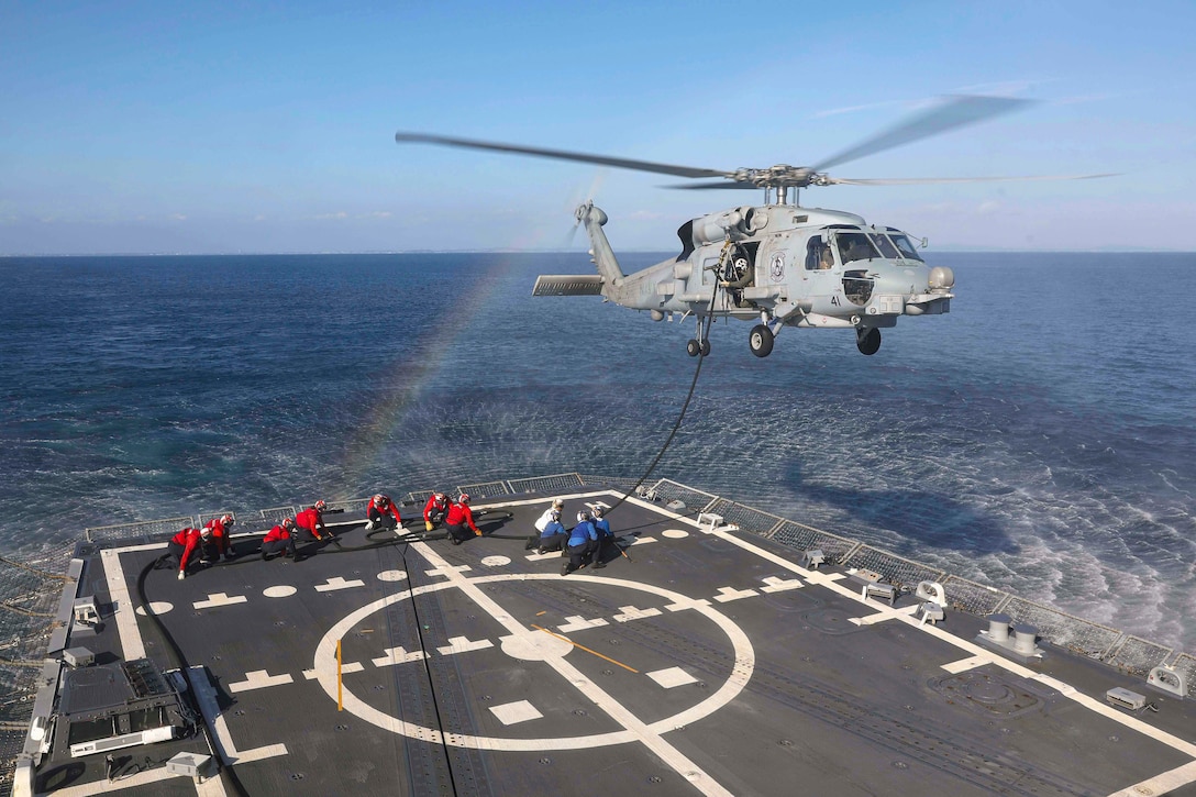 Sailors kneel on a ship at sea while holding a hose connected to a hovering helicopter with a rainbow to its left.