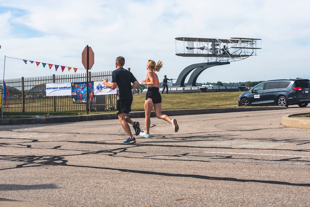 Two runners run past a model Wright-Flyer next to a military base gate.