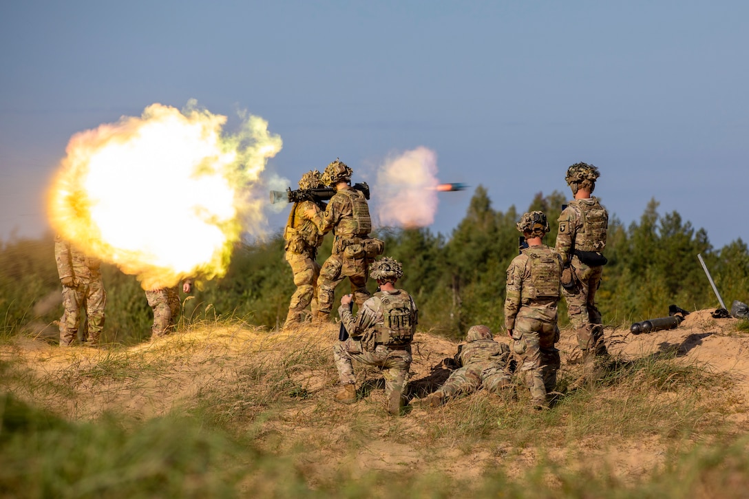 Soldiers stand or kneel as two fellow soldiers fire a weapon as an explosion arises to the left with a wooded area in the background.