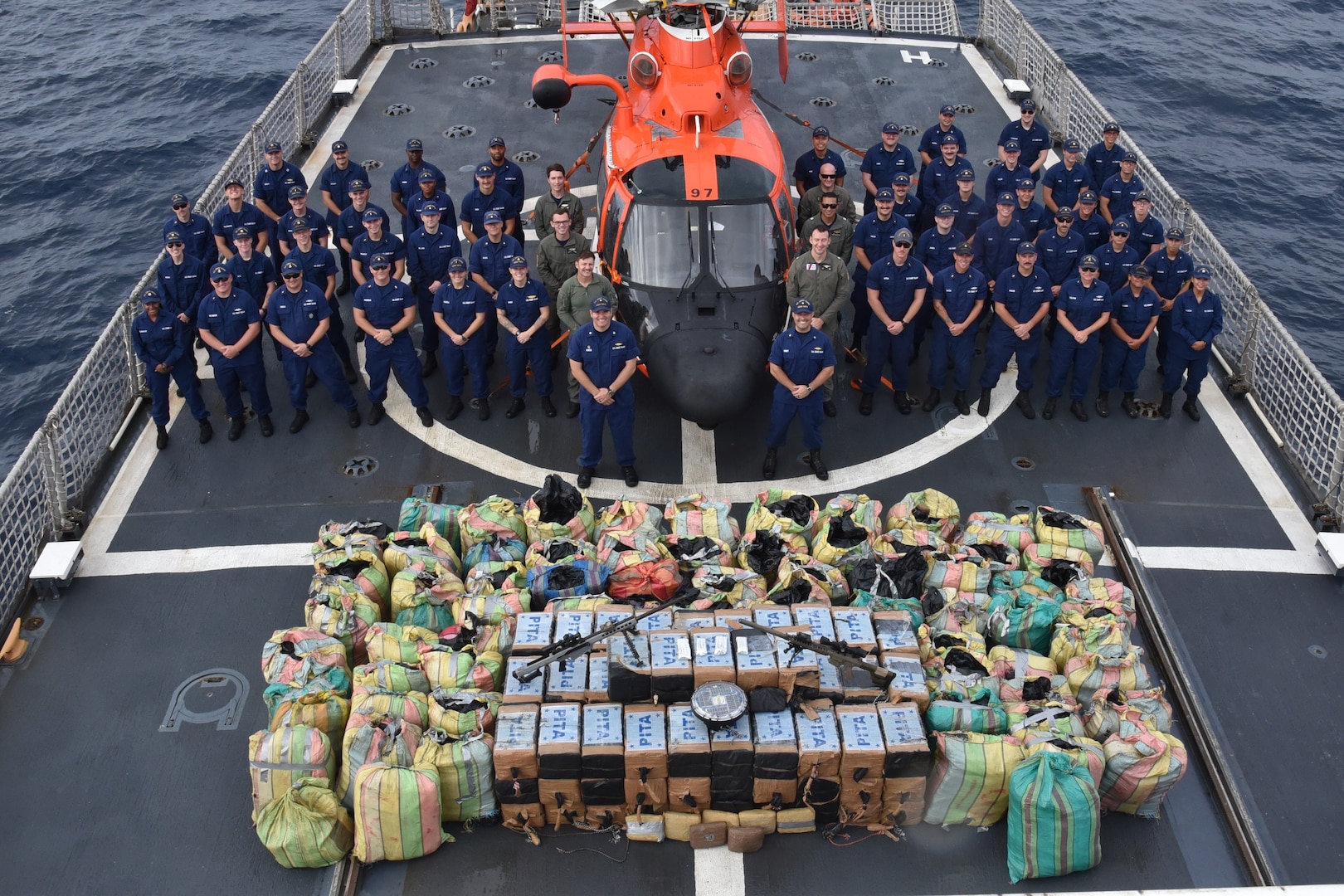 The crew of U.S. Coast Guard Cutter Thetis (WMEC 910) poses for a group photo on the flight deck with over 10,000 pounds of interdicted narcotics in the Eastern Pacific Ocean, Sept. 8, 2023. Thetis' crew patrolled the Eastern Pacific Ocean and Western Caribbean Sea in support of the Coast Guard’s mission to combat illegal drug trafficking. (U.S. Coast Guard photo by Ensign Lauren Daugherty)