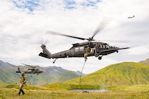 Naval Special Warfare operators fast-rope from two hovering helicopters into the mountains as an aircraft flies above.