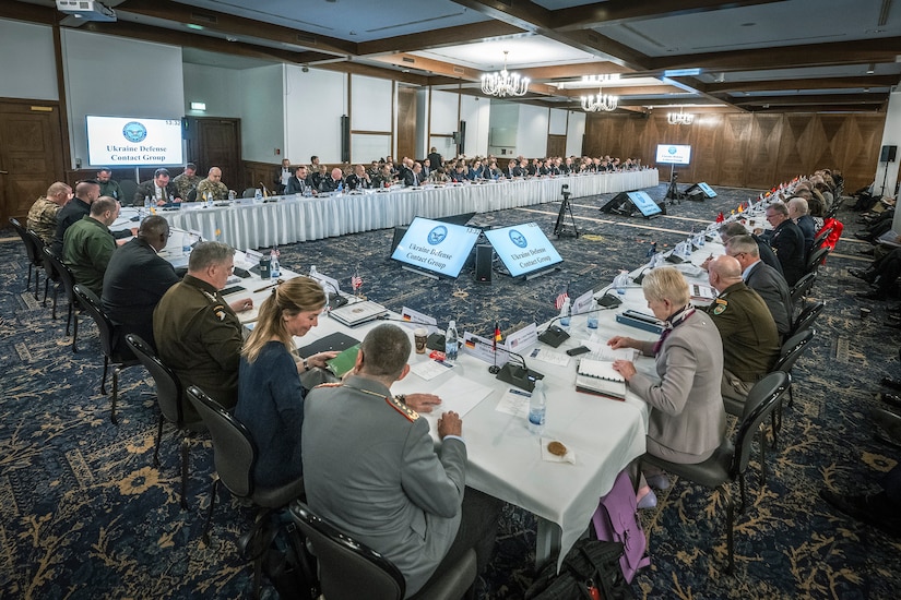 A large number of people are seated around a conference table. The sign indicates that they are attending a meeting of the Ukraine Defense Contact Group.