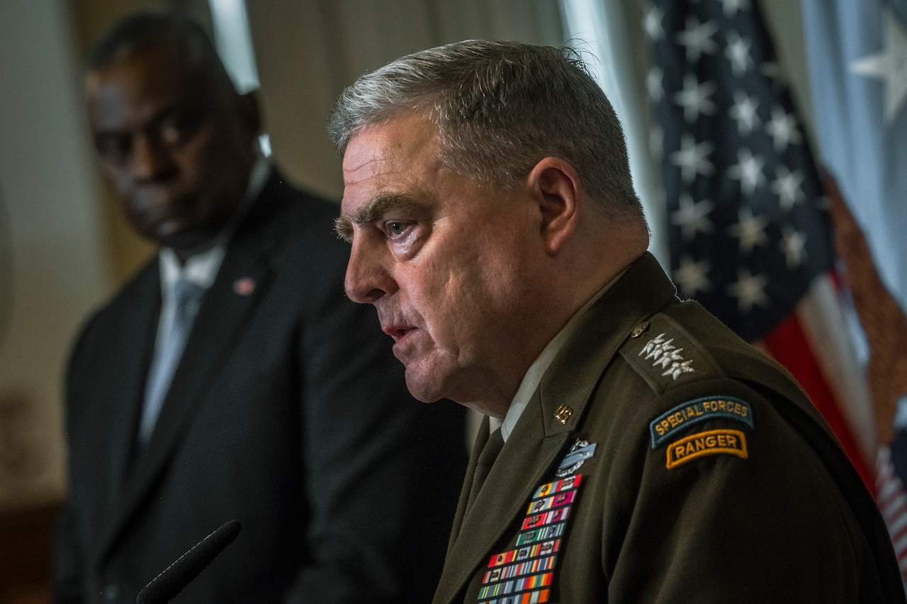 Two people, one in a military uniform and the other in a business suit, stand at lecterns.