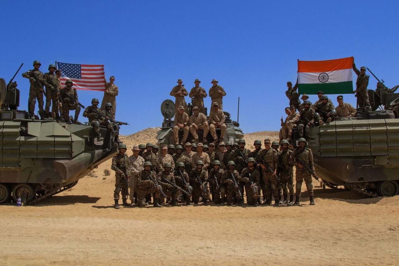 A group of troops pose with members of a foreign military as they hoist American and Indian flags while standing on armored vehicles.