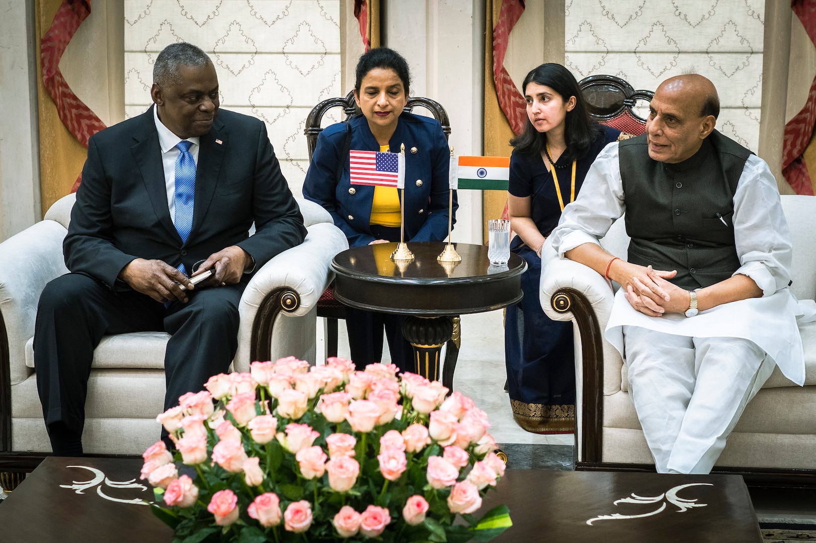 Two men in civilian attire are seated next to a table displaying American and Indian flags.