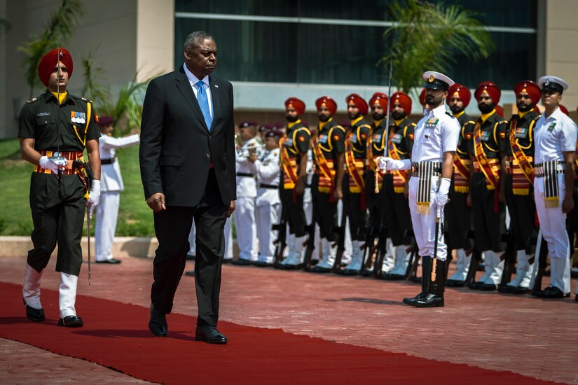 A man in civilian attire walks as members of a foreign military stand at attention.