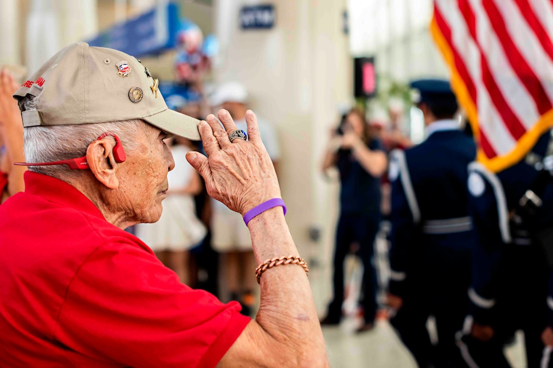 A man salutes an American flag as service members walk past in formation.