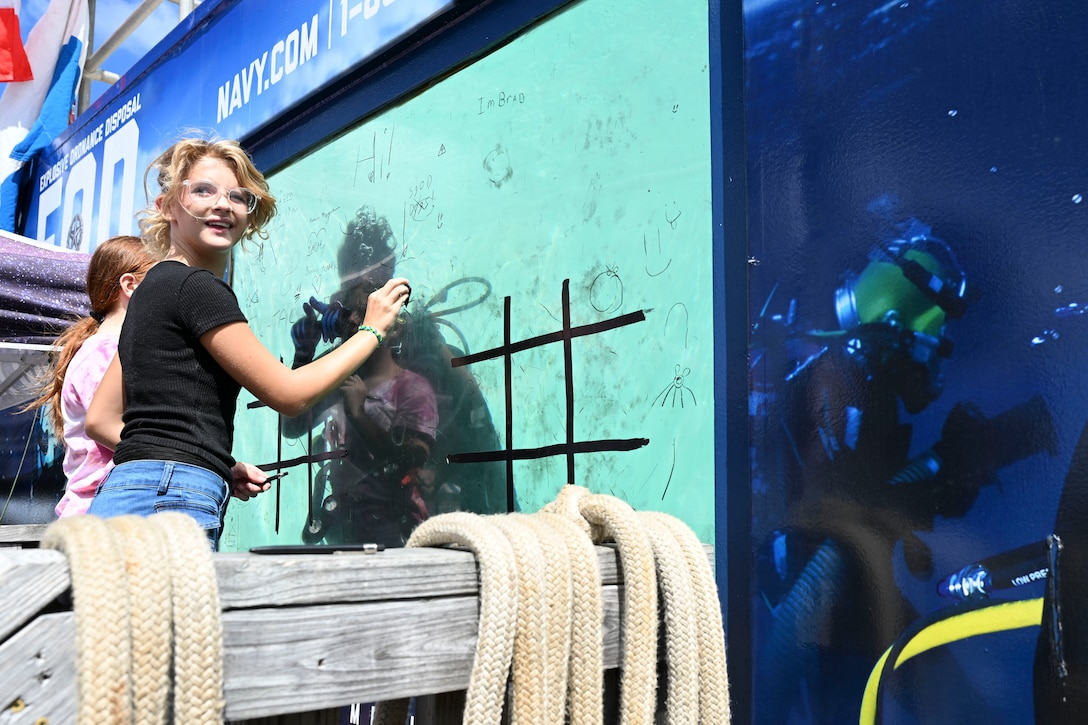 Children draw on a monitor while divers swim behind a glass screen.