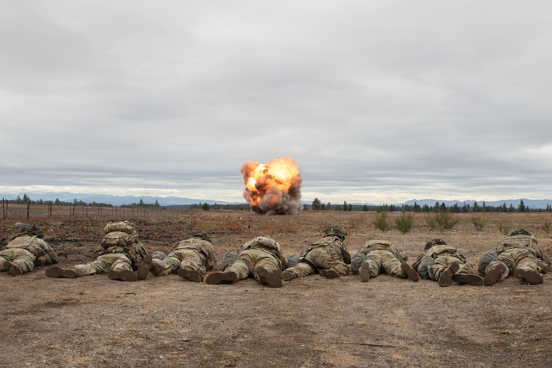 Soldiers lay on the ground as an explosion detonates in the distance.
