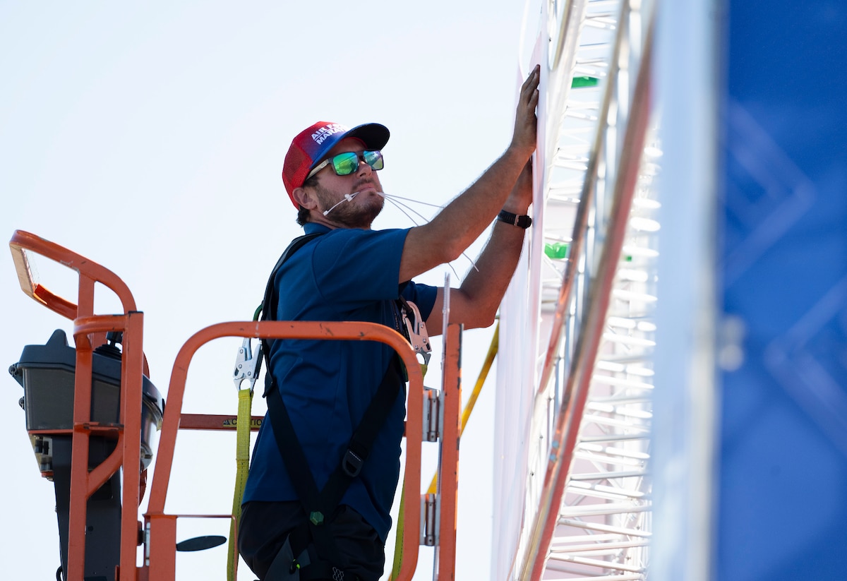 A man in a hat and sunglasses works raised in a bucket truck.