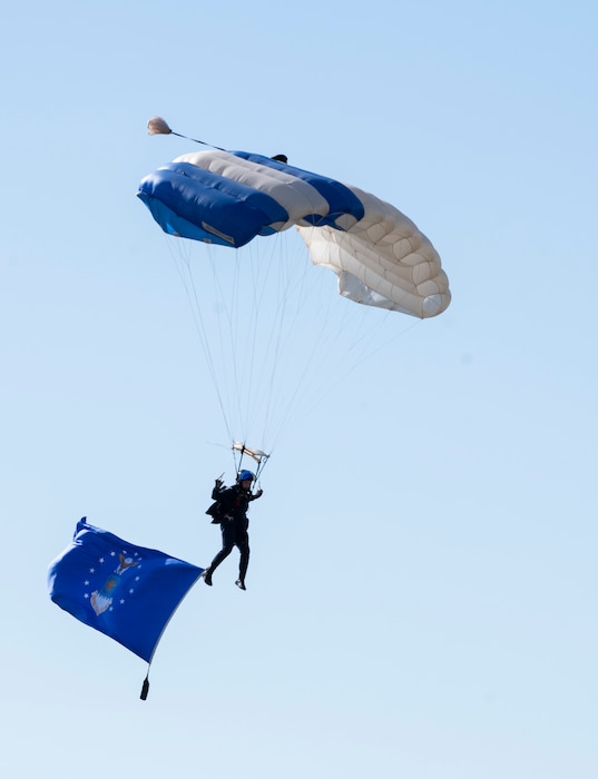 a man hangs from a square parachute. A large Air Force flag hangs below him.