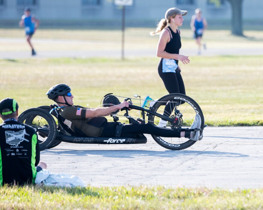 A man in a reclining  hand-crank bicycle passes a running woman.