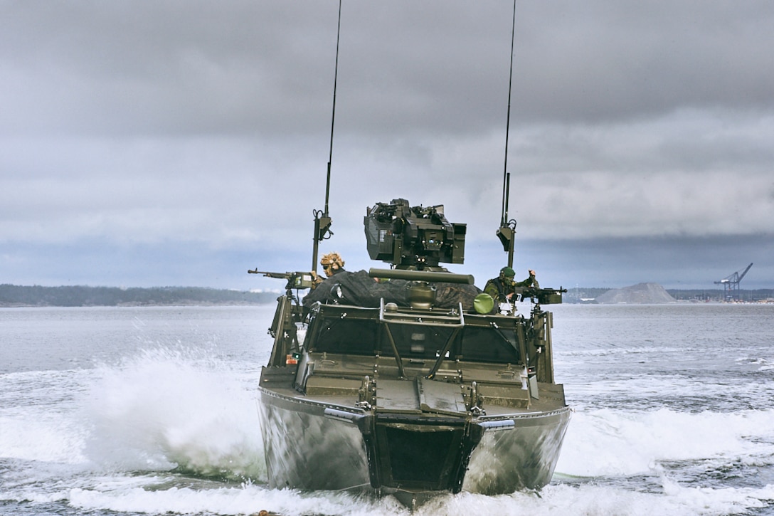 U.S. and Swedish marines aim weapons in opposite directions while transiting a body of water in a boat.