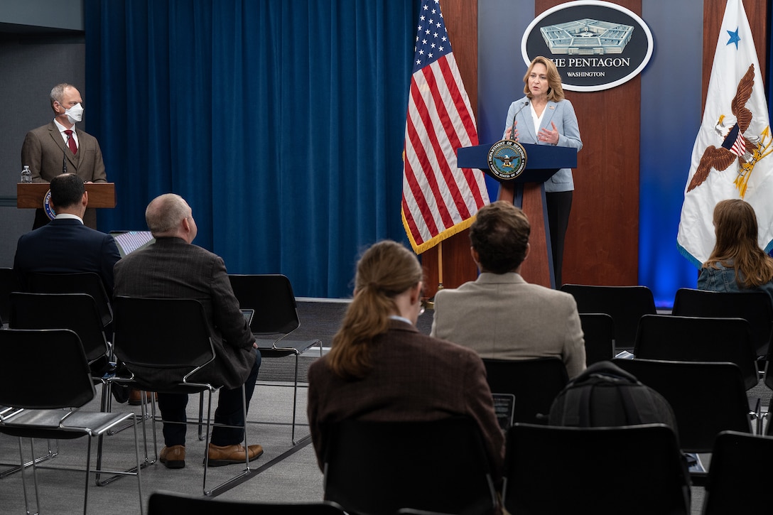 A person in business attire speaks from a lectern as others look on.