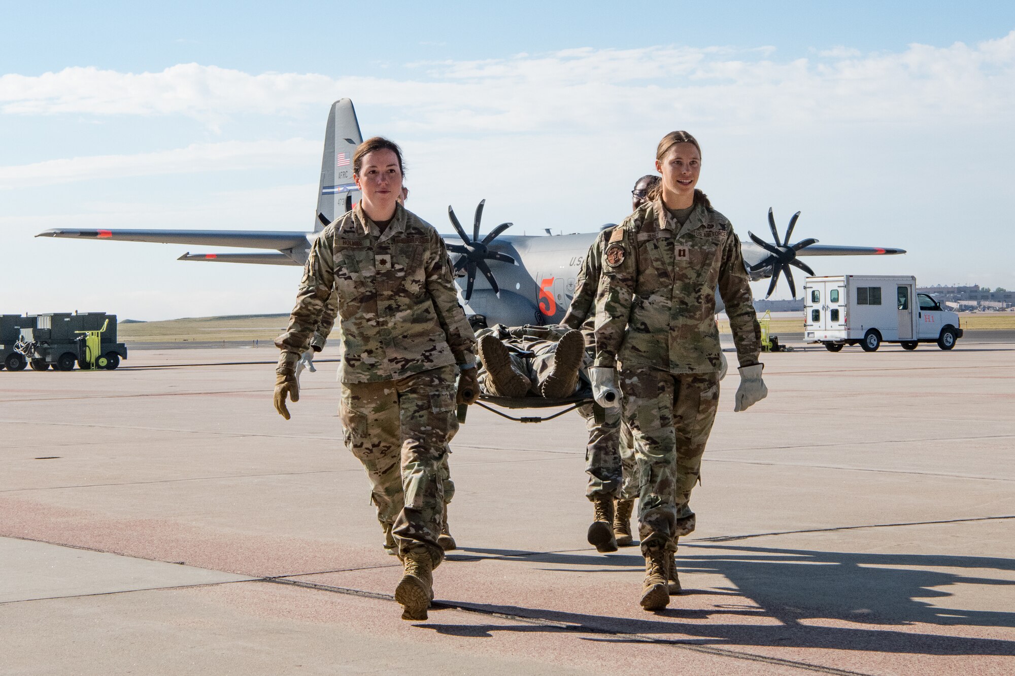 Four Airmen carry a patient in a litter with a cargo aircraft in the background on an aircraft parking ramp.