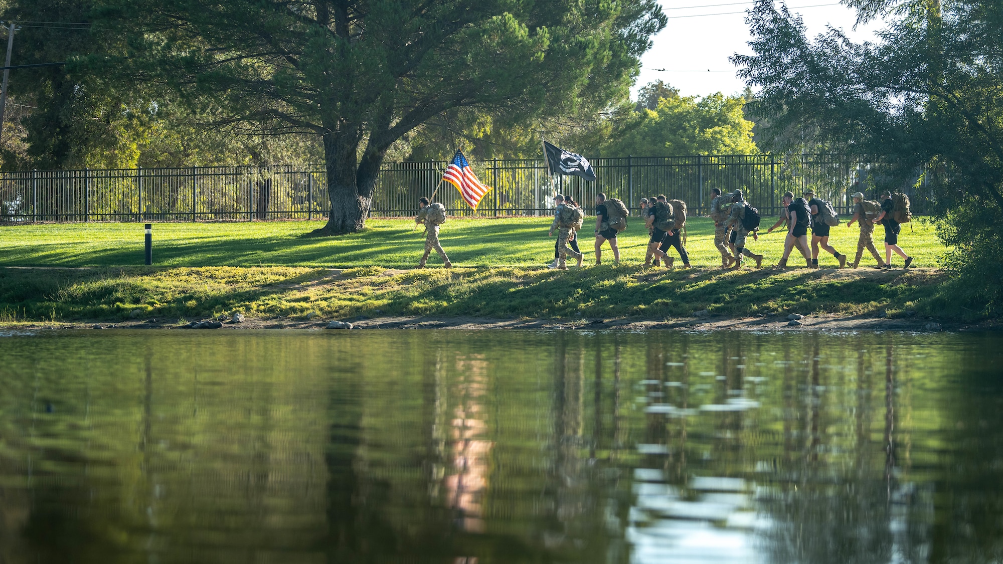 military members walk with flag around pond