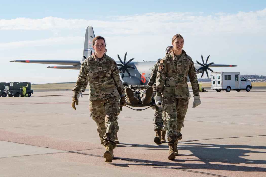 Four Airmen carry a patient in a litter with a cargo aircraft in the background on an aircraft parking ramp.