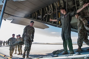 An Airman in a flight suit extends his arm toward an aircraft cargo bay to direct two Airman carrying a patient on a litter.
